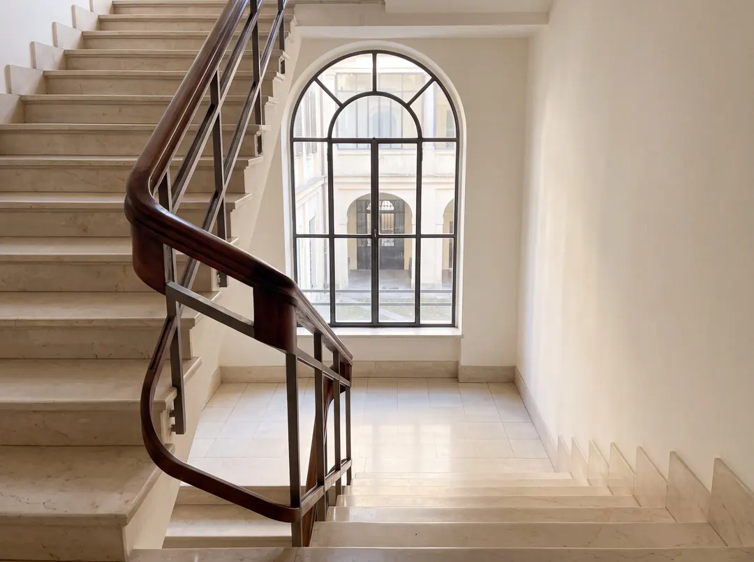 Scala condominiale con gradini in marmo puliti e corrimano in legno, luce da una finestra alta.